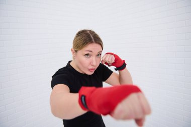 Güçlü, genç, sarışın bir kadının boks eldivenleriyle kameraya yumruk atarken, dövüş sanatı, kutu ve beyaz arka planda kickbox yaparken kendine güveni tamdır. Spor. Kardiyo çalışması. Dayanıklık