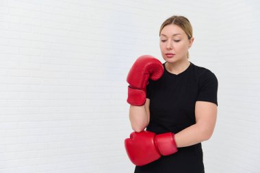 Beyaz, sarışın, 30 'lu yaşlarda, kırmızı boks eldiveni takan, reklam alanı için beyaz duvar arkasına izole edilmiş bir kadın. Atletik kadın boks antrenmanına hazır. Spor, disiplin, dayanıklılık.
