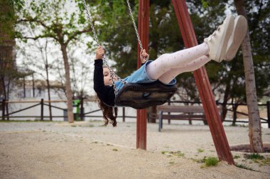 Kafkasyalı, rahat kot giyen, gülümseyen, dışarıda oyun alanında sallanırken rahatlatan sevimli kız çocuğu. - İnsanlar. Mutlu tasasız çocukluk kavramları. Yaşam biçimi