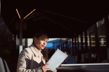 Paltolu bir kadın kafede güneşli bir kış gününün tadını çıkarıyor, elinde kitap tutuyor ve düşünüyor..