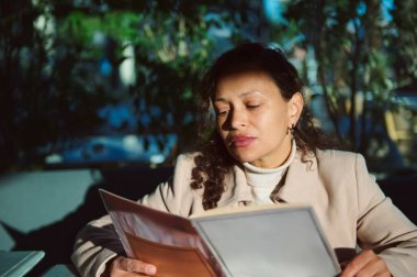 Açık bir kafede dikkatlice menü okuyan bir kadının portresi. Parlak güneş ışığı yüzünü ve çevresini aydınlatıyor, sakin ve odaklanmış bir atmosfer yaratıyor..