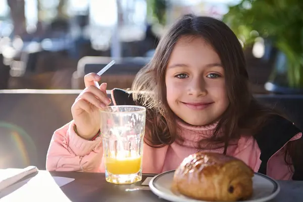 stock image A cheerful young girl enjoys a breakfast of orange juice and a pastry at an outdoor cafe. The peaceful morning atmosphere and her joyful expression create a warm and inviting scene.