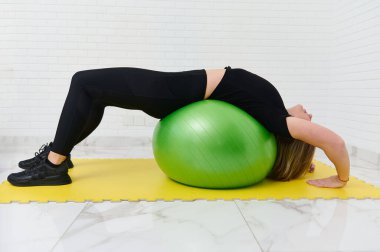 Genç sporcu bir bayan atlet ev stüdyosunda yeşil bir fitness topu üzerinde egzersiz yapıyor. Siyah atletizm kıyafetleri içinde sarı bir hasırın üzerinde esneyerek güç ve esnekliğe odaklanıyor..