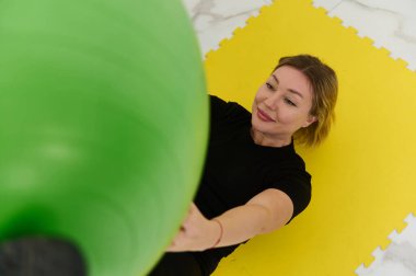 Genç sporcu bir bayan sporcu evde sarı bir paspasın üzerinde yatarken yeşil bir fitness topuyla egzersiz yapıyor. Odaklanmış ve kararlı, rahat bir ev stüdyosunda fitness rutinini sürdürmekten hoşlanıyor..