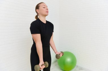 Genç atletik kadın evde fitness topuyla egzersiz yapıyor. Odaklanmayı ve dengeyi koruyor, ev stüdyosunda kişisel fitness hedeflerine bağlılığını ve bağlılığını gösteriyor..