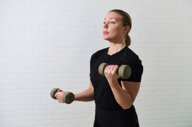 Genç bir bayan atlet evde halterlerle çalışıyor. Görüntü onu odaklanmış ve kararlı bir ruh halinde, iyi ışıklandırılmış bir mekanda spor yaparken gösteriyor..