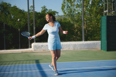 40 'lı yaşlarda bir kadın açık havada güneşli havada tenis oynarken görülüyor. Spor kıyafetleri içinde ve oyundan zevk alıyor gibi görünüyor, bir eğlence ve uygunluk duygusu aktarıyor..