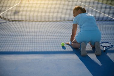 40 'lı yaşlarda bir kadın dışarıda tenis oynuyor, diz çöküp tenis topuna uzanıyor. Raketi yanında yatarken odaklanmış ve kararlı görünüyor. Sakin güneşli hava.