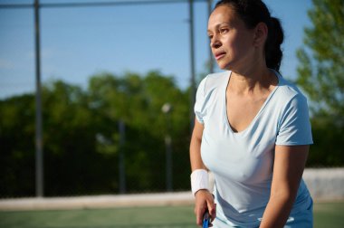 Açık havada güneşli gökyüzünün altında tenis oynayan orta yaşlı bir kadın. Yoğun odaklanmış ve topa vurmaya hazır, açık mavi bir spor kıyafeti ve bilekliği var..