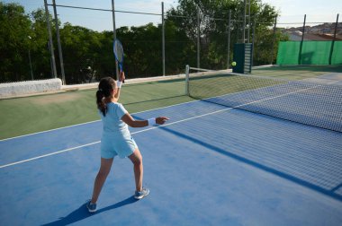 40 'lı yaşlarda bir kadın açık hava kortunda aktif olarak tenis oynuyor. Raketiyle topa vurmaya hazırlanıyor, atletizm ve coşku gösterisi yapıyor..