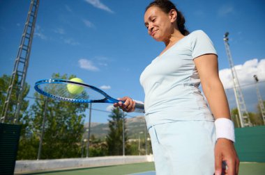 40 'lı yaşlarda dışarıda tenis oynayan neşeli bir kadın. O bir tenis raketi tutuyor ve topa odaklanmış ve kendine güvenerek bakıyor. Parlak, güneşli bir gün spor atmosferini güçlendiriyor..