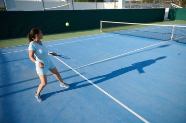 40 'lı yaşlarda bir kadın açık hava kortunda aktif olarak tenis oynuyor. Görüntü, topa vururken odaklanmasını ve atletizmini yakalıyor. Açık ve güneşli bir günde sporun zevkini vurguluyor..