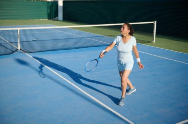40 'lı yaşlarda parlak mavi bir kortta tenis oynayan odaklanmış bir kadın. O, azim ve beceri göstererek sporla meşgul. Bu resimde etkin bir yaşam tarzı ve açık hava etkinliği vurgulanmıştır.