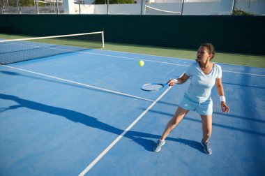 40 'lı yaşlarda bir kadın açık hava kortunda aktif olarak tenis oynuyor. Salıncağın ortasında, tenis topuna vurmaya odaklanmış. Güneşli hava dalgalı mavi saray yüzeyini vurguluyor.