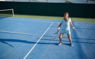 Spor kıyafetleri içinde elinde tenis raketi tutan orta yaşlı bir kadın. Dikkatle maça odaklanmış. Sahne güneşli bir günde açık tenis kortunda geçiyor. Kararlılık ve atletizm gösteriliyor..