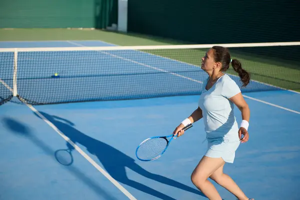 Stock image Woman in her 40s playing tennis on an outdoor court. She is in mid-action, hitting the ball with focus and determination. The clear blue sky and green surroundings add a vibrant atmosphere.