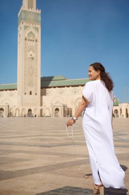 Aydınlık ve güneşli bir günde uzun bir minareyle karışık bir caminin önünde durmuş dua boncukları tutan geleneksel beyaz elbiseli bir kadın huzur ve maneviyat yayıyor..