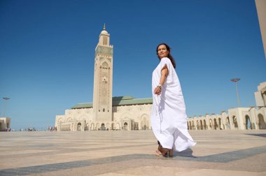 Geleneksel elbiseli kadın, Kazablanka, Fas 'taki Hassan II Camii' nin önünde güven içinde duruyor. Açık mavi gökyüzü ve caminin mimari güzelliği huzurlu ve görkemli bir manzara yaratır..