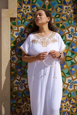 Geleneksel beyaz elbiseli bir kadın canlı, renkli desenli bir duvarın önünde tespih ile dua ederken görülüyor. Huzurlu ve huzurlu görünüyor, ruhani bir anı kucaklıyor..