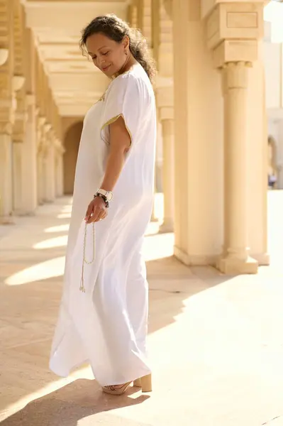 stock image Elegant woman in flowing white dress walking gracefully through sunlit corridor with beautiful architectural details. Serene and contemplative atmosphere is enhanced by the soft light, surroundings.