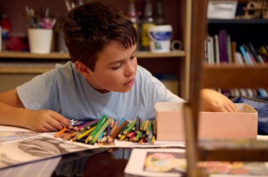 Genç çocuk renkli kalemlerle dolu bir resim sınıfında dikkatlice resim çiziyor. Sınıf çevresi yaratıcılığı ve öğrenmeyi teşvik eder..