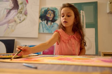 Genç bir kız resim dersi sırasında boya fırçası kullanarak resim yapıyor. Renkli bir atölyede yaratıcılık ve konsantrasyonu vurguluyor..