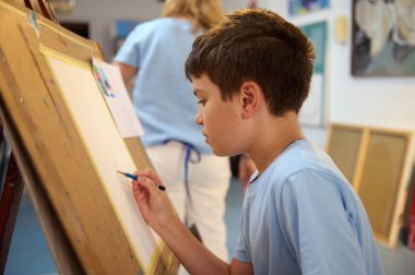 Resim dersinde çizimlerine konsantre olan genç bir çocuk atölye ortamında bir öğretmen tarafından rehberlik ediliyor. Çocuklar için yaratıcı ve eğitici etkinlik.