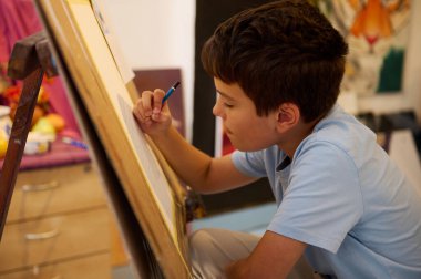 Resim dersinde bir öğretmenin rehberliğinde diğer çocuklarla birlikte çizimlerine konsantre olan genç bir çocuk. Atölyede çeşitli sanat malzemeleri ve yaratıcı bir ortam yer alıyor..