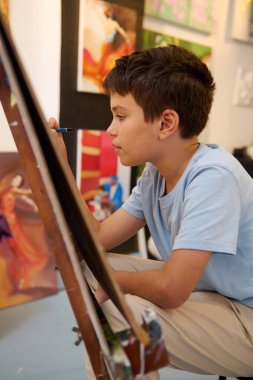 Sanat dersi sırasında tuvale dikkatlice resim yapan genç bir çocuk. Arkaplanda renkli sanat eserleriyle çevrili yaratıcı sürecine derinden dalmış durumda..