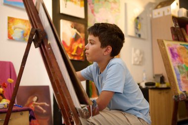 Sanat dersi sırasında dikkatle resim yapan genç bir çocuk. Sınıf çeşitli bitirilmiş sanat eserlerini sergiliyor, yaratıcılık ve öğrenme için ilham verici bir ortam yaratıyor..