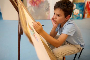 Çocuklar resim dersi alıyor, resim yapıyor, atölyede öğretmenle resim yapıyor. Genç çocuk, yaratıcılık ve konsantrasyon sergileyerek sanatına derinden odaklanmış durumda.