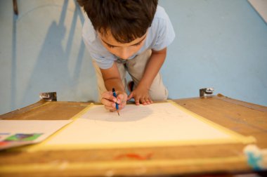 Genç bir çocuk resim dersinde resim yapmaya dalmış. Çocuk sanat eserlerinin üzerine eğiliyor, bir atölye ortamında şaheserini yaratmaya odaklanıyor..
