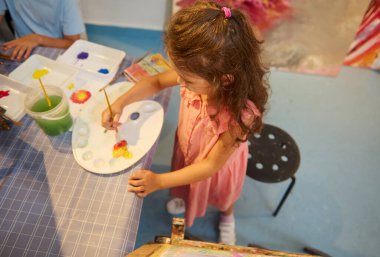 Çocuklar resim dersi alıyor, resim yapıyor, atölyede öğretmenle resim yapıyor. Sanat malzemelerinin olduğu bir masada resim yapan genç bir kız sınıf arkadaşlarıyla yaratıcı aktivitelerden zevk alıyor, yaratıcılığı teşvik ediyor ve öğrenmeyi teşvik ediyor..
