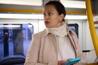 Paltolu ve atkılı bir kadın otobüste giderken akıllı telefon kullanıyor. O, düşünceli bir şekilde pencereden dışarı bakar..