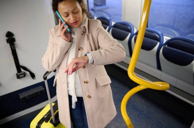Trende telefonla konuşan ve saatini kontrol eden bir kadın. Sarı bir bavulla seyahat ediyor gibi görünüyor. Sahne aciliyet ve modern seyahat içeriyor..