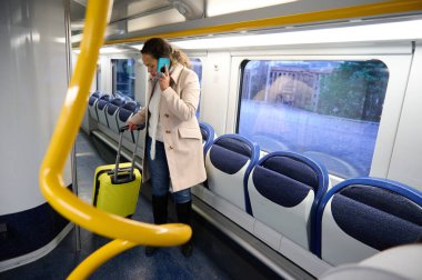 Bavulunu tutan ve trende seyahat ederken telefonla konuşan bej ceketli bir kadın. Tren neredeyse boş, ve arka plan şehir ortamını gösteriyor..