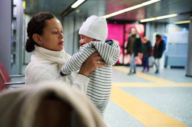 Arka planda insanlar yürürken bebeğini kapalı bir alanda tutan bir anne. Anne bebeğe sevgiyle bakar..