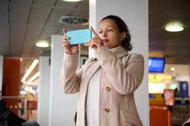 Havaalanı terminalinde fotoğraf çekmek için bej ceketli bir kadın akıllı telefonunu kullanıyor. Arka planda diğer gezginlerle birlikte parlak ve modern iç mekan.