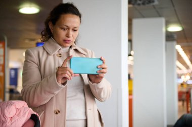 Bej ceketli bir kadın kapalı bir alanda fotoğraf çekmek için akıllı telefonunu kullanıyor. O anı yakalamak için odaklanmış ve kasıtlı görünüyor..