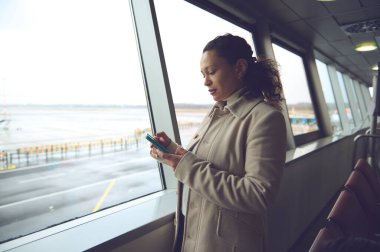 İş kadını havaalanında uçağını beklerken telefonunu kontrol ediyor. Piste bakan büyük pencerelerden gelen doğal ışık.