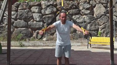 Sporcu güneşli bir günde parkta göğüs egzersizleri yapar. Uzaklaştırma eğitim sistemi kullanıyor, arka planda sarı bir bank. Bu video fitness, sağlık ve açık hava egzersizi temaları çağrıştırıyor.
