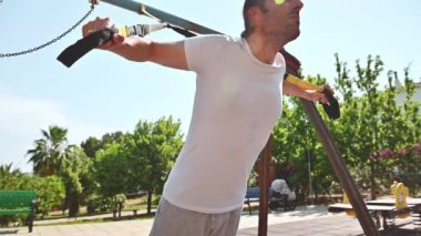 Formda bir adam güneşli bir günde parkta askılarla egzersiz yapar. Formunu geliştirmeye odaklanmış ve dışarıda çalışmaktan zevk alıyor..