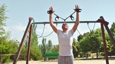 Güneşli bir günde 40 'lı yaşlarında bir adam parkta spor yapıyor. Kas yapmak için süspansiyon kayışları kullanıyor. Odaklanmış ve kararlı..