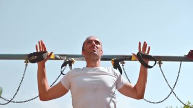 Alnında ter olan motive olmuş bir adam spor salonunda spor yaparak vücudunun üst kısmını çalıştırıyor. Kontrollü hareketlerle kendini yukarı çeker. Gücünü ve uygunluğunu gösterir..