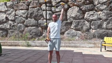 Kırklı yaşlarda, kısa sakallı, beyaz tişörtlü ve gri şortlu bir adam açık hava spor salonunda spor yapıyor. TRX direniş bantları kullanıyor ve spor yaparken gülümsüyor..