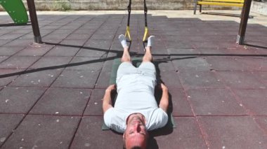 Formda bir adam güneşli bir günde açık havada egzersiz minderinde yatar. O, bacak egzersizi yapmak için fitness kayışları kullanıyor. Egzersiz rutinine odaklanmış..