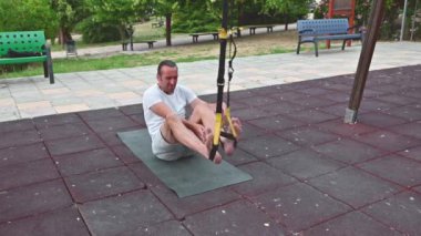 30 'lu yaşlarda, açık hava spor salonunda TRX süspansiyon eğitimi alan fit bir adam. Yerde yatarak ve ayaklarını kayışlarla yukarı çekerek vücut ağırlığı egzersizi yapıyor..