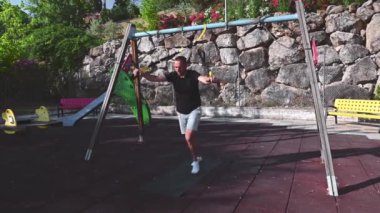 Formda bir adam güneşli bir günde parkta süspansiyon eğitim sistemini kullanarak egzersiz yapar. Kolları havada, bacakları ve üst vücudu üzerinde hareket ediyor. Sınırlarını zorluyor..