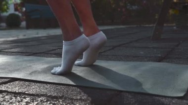 Beyaz çoraplı bir kadının ayaklarını kapayarak parkta spor yapmak için ısınıyor. Yoga minderinin üzerinde duruyor ve baldır kaldırıyor..