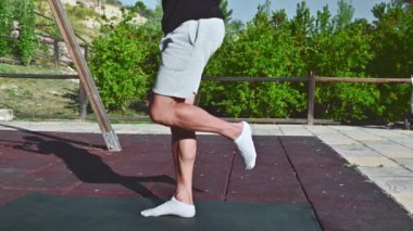 Gri şortlu ve siyah tişörtlü bir adam açık hava parkında siyah bir paspas üzerinde bacak egzersizi yapıyor. Isınıyor, bacaklarını açıyor ve antrenman için hazırlanıyor..